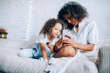 Sevimli küçük kız evde annesinin kanepesinde uzanıp birlikte akıllı telefon kullanıyor ve boş vakitlerinde vakit geçiriyor.