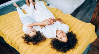 Büyüleyici etnik kadın ve aynı tatlı kız el ele tutuşurken yatakta dinleniyor ve kameraya gülümsüyor.