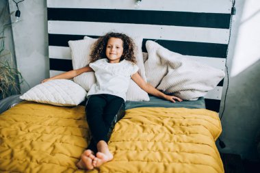 Bukleleri olan, ev kıyafetleri giyen, yastıkları olan ve kameraya gülümseyen sevimli küçük bir kız.