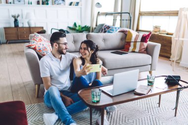 Mutlu bir çift kot pantolon giyer ve rahat koltukta kucaklaşır. Dairede modern iç mekan ile selfie çeker ve birbirlerine bakarlar.