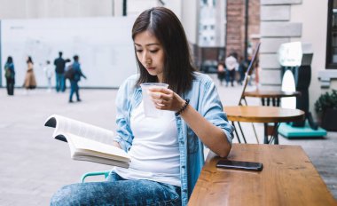 Esmer, güzel, Asyalı bir kadın soğuk, ferahlatıcı içecekler içip kitap okurken ahşap masada, akıllı telefonlu bir kafede oturuyor.