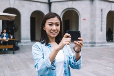 Neşeli, güzel Asyalı kadın akıllı telefonuyla fotoğraf çekiyor ve arka planda bulanık bir şekilde sokağın ortasında kameraya bakıyor.
