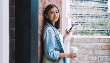 Tuğla duvara yaslanan ve telefon kullanırken kameraya bakan gündelik giysiler içinde çekici bir etnik kadın.