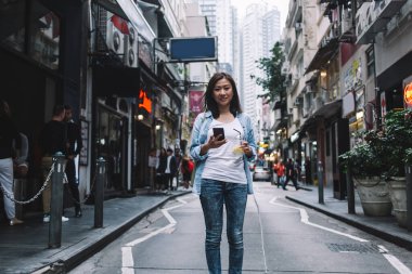 Neşeli, güzel, yetişkin, sıradan kıyafetler içinde Asyalı bir kadın. Canlı bir caddenin ortasında cep telefonuyla duruyor. Plastik bardakta içkisini tazeliyor ve kameraya bakıyor.