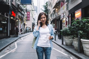 Neşeli, güzel, yetişkin Asyalı kadın, gündelik kıyafetleriyle caddenin ortasında cep telefonundan arıyor, sıcak içeceği tek kullanımlık bardakta.