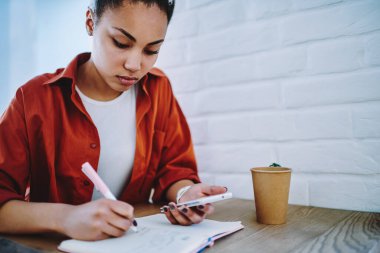 Üniversite sınavına hazırlanan ciddi bir kız öğrenci eğitim için ders kitabına cevaplar yazarken, cep telefonuyla ders için bilgi arayan koyu tenli genç hippi kız.