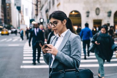 Gözlüklü saygıdeğer bir bayan girişimcinin elinde moda çantasıyla yol kenarında durması ve bulanık arka planda sosyal ağda mesajlaşması