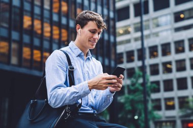 Mavi gömlekli, siyah çantalı, kablosuz kulaklıklı, New York 'ta oturan ve akıllı telefondan mesaj atan memnun bir adam.