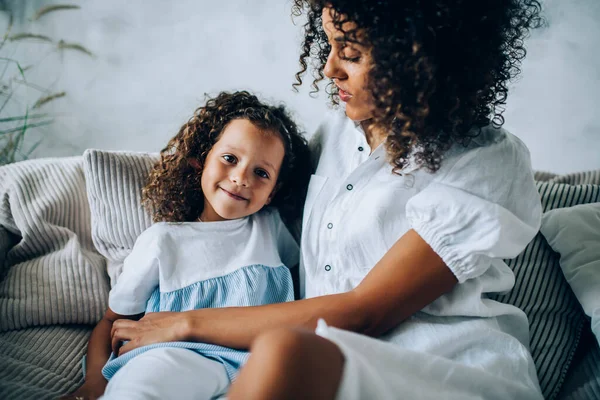 Crop ethnic woman with cute little girl sitting on comfy sofa at home embracing with love and spending time together