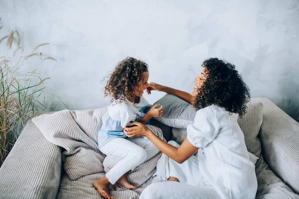 Side view of happy beautiful woman and girl both with curly hair having fun at home while chilling on comfy sofa