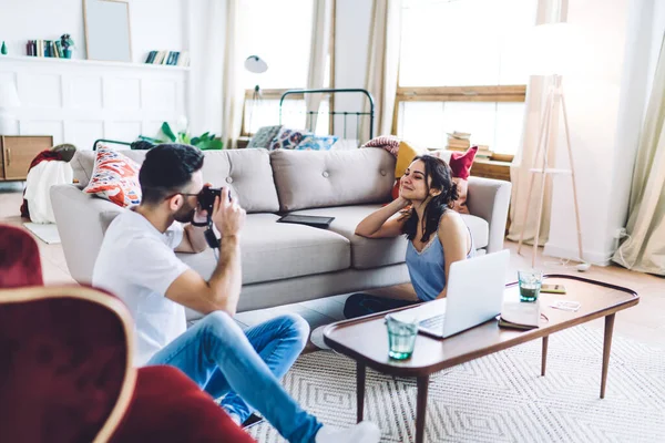 Millennial couple sitting on floor between sofa and coffee table with laptop while boyfriend holding photo camera and taking picture of girlfriend