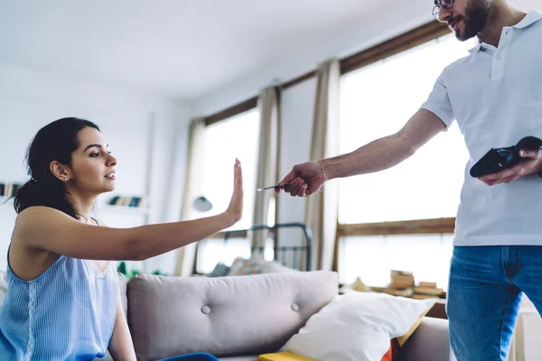 Side view of smart confident woman gesturing with stretched hand sitting on sofa and refusing credit card suggesting by crop man