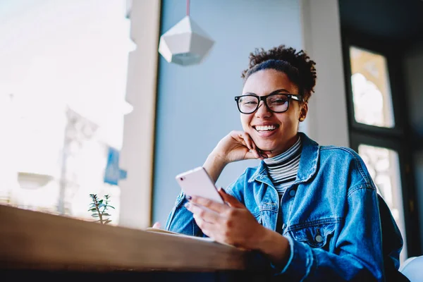 Portrait of excited female user laughing from joke read on web page via smartphone gadget and looking at camera during free time,cheerful dark skinned woman in spectacles browsing internet on cellular