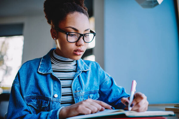 Attractive woman in eyeglasses for vision correction writing essay in notepad concentrated on ideas, serious african female journalist dressed in jeans jacket notes questions for next interview