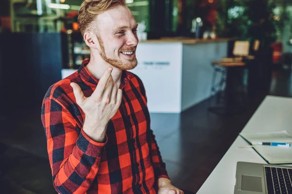 Positive hipster guy in stylish outfit laughing indoors during leisure time with laptop computer, happy young man smiling during break from planning week schedule via textbook and modern netbook