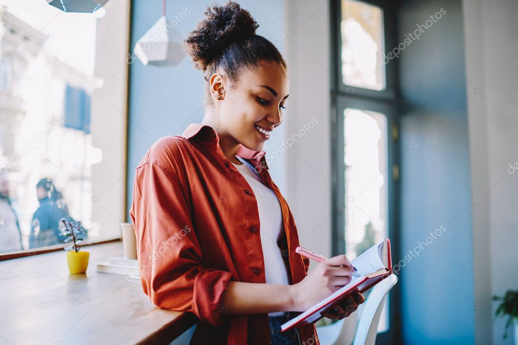 Happy dark skinned female student enjoying time for autodidact writing essay with excited idea in textbook, cheerful woman notes pleasant moments from day in notebook during free time in cafeteria