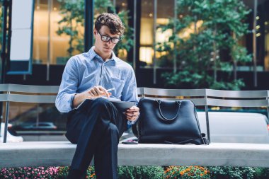 Gözlüklü yakışıklı genç bir erkek ofis binasının önündeki bankta oturuyor Manhattan, New York 'ta bir sokakta tablet okuyor.
