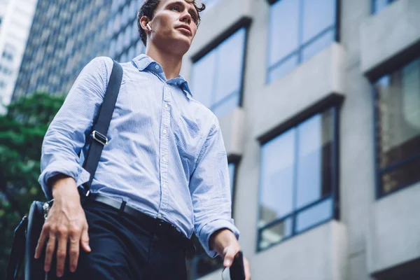 From below of confident young man in classic outfit holding leather bag having phone call by wireless headphone while walking on street beside of commercial buildings of New York city