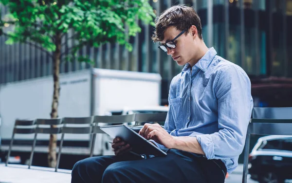Confident man entrepreneur chatting on digital tablet with client while sitting on bench at urban setting, intelligent male lawyer in formal wear reading electronic book during work break in city