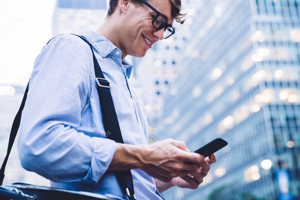 Low angle side view of male employee in shirt and glasses chatting on phone on background of New York smiling