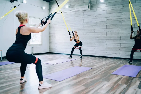 Couple of women with blonde hair in sport clothes with resistance band on knees squatting with rubber expander near fitness mats in gym