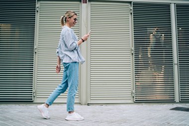 Şehrin sokaklarında yürürken günlük giysiler içinde gülümseyen ve akıllı telefonlara bakan neşeli genç bayanın yan görüntüsü.