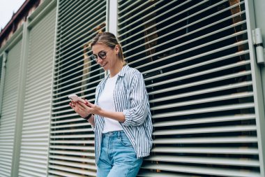 Penceresi kör binaya yaslanan ve gülümseyen ve rahatlatıcı bir şekilde akıllı telefon kullanan hoş bir bayan.