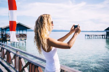 İnce, bronz tenli, beyaz bluzlu, güneş gözlüklü genç bir kadın. Denizde ahşap iskelede duruyor ve gün ışığında bulanık arka planda fotoğraf çekiyor.