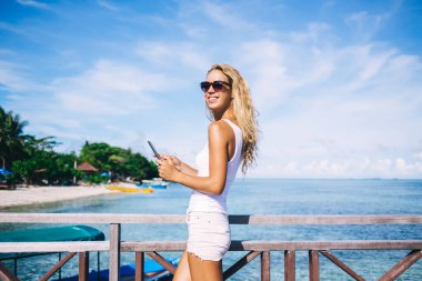 Beyaz bluzlu, güneş yanığı bir kadın, şort ve güneş gözlüğü takıyor. Deniz kenarındaki ahşap korkuluklarda duruyor ve gün ışığında akıllı telefonda daktilo yazıyor.