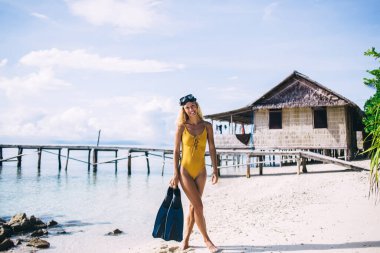 Gözlüklü güzel sarışın, gülümseyen ve palet taşıyan. Sahilde yürürken kabin ve bulutlu iskelenin arka planında.