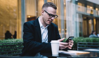 Siyah ceketli ve gözlüklü zeki adam akıllı telefonuyla mesajlaşırken bir fincan kahveyle kaldırımdaki masada oturuyor.