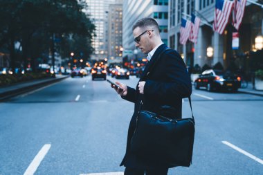 Siyah iş ceketli, omuz çantalı gözlüklü, akıllı telefon kullanan ve büyük şehir yolunu geçen bir adamın yan görüntüsü.
