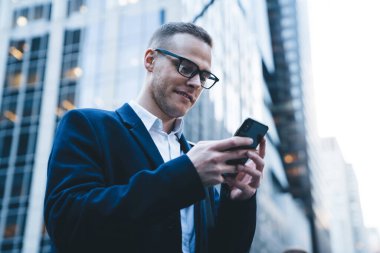 Palto ve gözlükleriyle gülümseyen genç ve yakışıklı girişimci şehir arka planında cep telefonu kullanırken