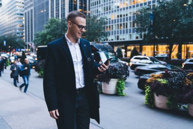 Şehir merkezindeki kalabalık bir caddenin ortasında resmi giyinmiş genç ve yakışıklı girişimci akıllı telefondan bilgi okurken gülümsüyor.