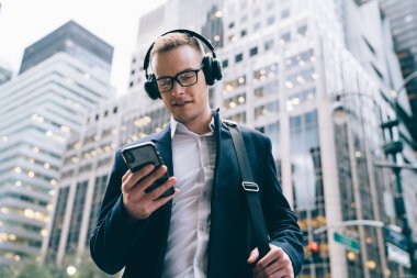 Gözlüklü, neşeli, yakışıklı, takım elbiseli, takım elbiseli ve kablosuz kulaklıkla müzik dinleyen bir işçiden. Ticari binaların bulanık arka planında cep telefonlarını tararken. 