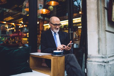 Yaşlı sakallı, gözlüklü ve takım elbiseli bir adam caddedeki kafede akıllı telefondan mesajlaşırken bacak bacak bacak üstüne atmış bir şekilde oturuyor.