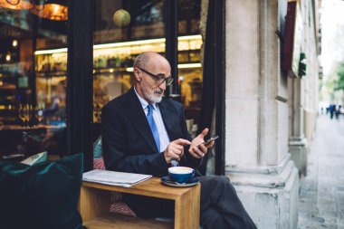Gözlüklü ve takım elbiseli yaşlı bir adamın camlara yaslanıp sokakta kahve fincanıyla masanın yanındaki akıllı telefona mesaj atması.
