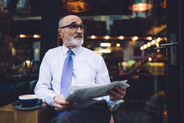 Gözlüklü, beyaz tişörtlü, bacaklarını çapraz yapıp caddede cama bakan yaşlı bir adam.