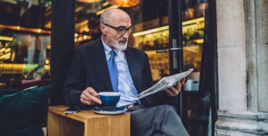 Yaşlı odaklı sakallı bir erkek, bardaklı kahve fincanı ve takım elbiseli, bacak bacak üstüne atmış ve pencerelere gazete okuyor.