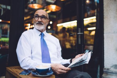 Gözlüklü ve beyaz tişörtlü kıdemli sakallı işadamı kahve fincanı ile masanın yanında oturuyor ve caddenin camına bakıyor.