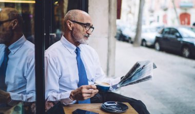 Beyaz tişörtlü, kahve fincanı olan, camların ve yolların yakınında oturan ve gazete okuyan kıdemli ciddi bir kıdemli adam.