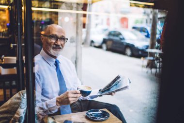 Pencereden, beyaz tişörtlü, kahve fincanı ve gazete kupasıyla şehrin caddesine bakan yaşlı, kısa boylu, kısa saçlı bir adam.