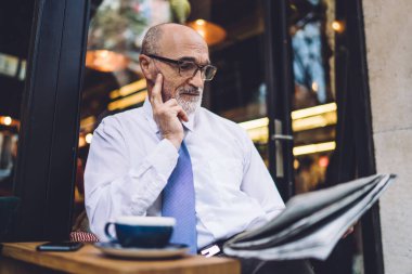 Yaşlı, kısa saçlı, beyaz gömlekli, kahve fincanı olan bir adam şehrin pencerelerine dokunup gazete okuyor.
