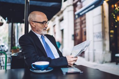 Gözlüklü başarılı yaşlı adam kaldırımdaki kafede bir fincan kahve ve akıllı telefonla masada oturuyor ve endişeli bir şekilde gazete okuyor.