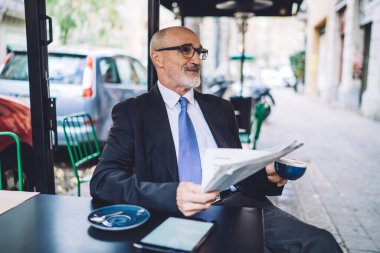 Caddedeki kafede ofis elbisesi ve mavi kravat takan neşeli son sınıf öğrencisi. Üzerinde kahve fincanı ve gazete var.