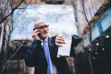 Olgun, kendine güvenen, iyi giyimli, elinde gazete olan beyaz saçlı, akıllı telefondan konuşan ve binanın dışındaki kol saatine bakan bir adamdan.