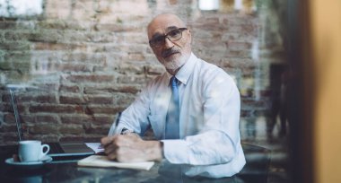 Gri saçlı, gözlüklü, kel bir adamın resmi kıyafetlerini incele. Not defterine not al. Kafede oturup kameraya bakarken dizüstü bilgisayarını kullan.