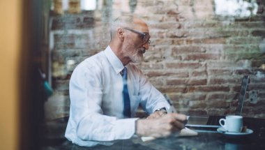 Gözlüklü, gri saçlı bir adamın kafeteryada dizüstü bilgisayarla otururken not alması.