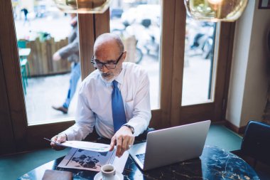 Yukarıdan, gözlüklü, kel, resmi giysiler giymiş, laptopun önünde oturmuş doküman okuyan, kafede oturan yaşlı bir adam.