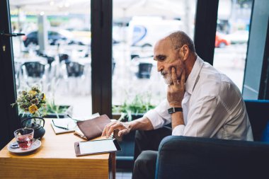 Kafede çay ve not defteriyle masada otururken beyaz gömlekli yaşlı bir adamın arka plan penceresine elini koyuşunun yan görüntüsü. 